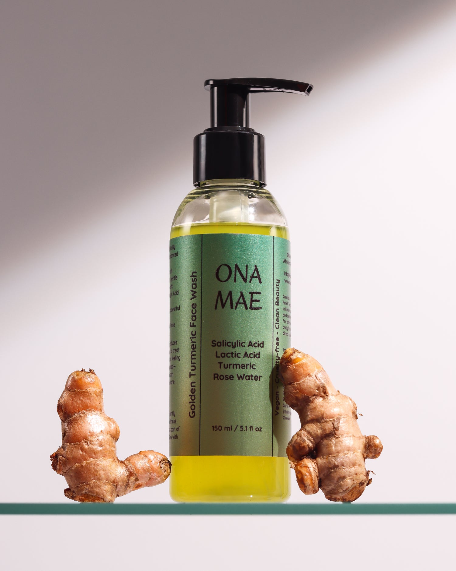 A 100 ml pump bottle of ONA MAE Golden Turmeric Face Wash, filled with yellow liquid, displayed on a glass shelf with two fresh turmeric root pieces on either side against a neutral gray background.