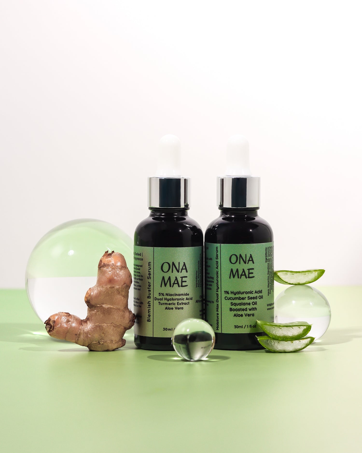 Two dark amber dropper bottles labeled ONA MAE stand on a pale green surface against a white background, styled with a ginger root, aloe slices, and clear glass spheres.